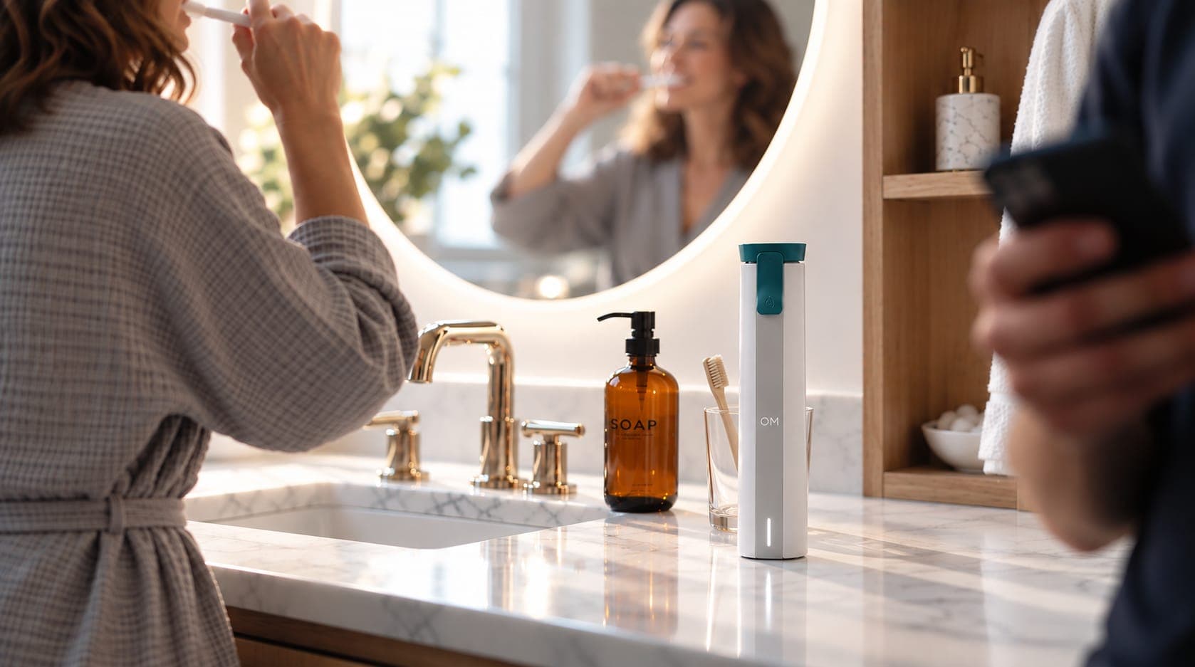 Le lecteur OM sur un comptoir de salle de bain en marbre ensoleillé, une femme se brossant les dents au miroir à côté et une personne consultant son téléphone au premier plan.
